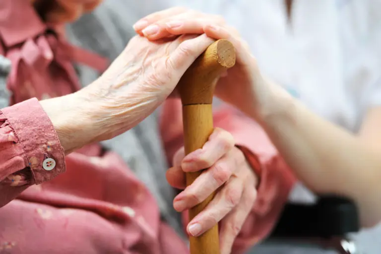 Pflegekraft hält unterstützend die Hand einer Seniorin mit Gehstock.